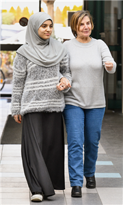 Eine Seniorin und eine junge Frau mit Kopftuch gehen Hand in Hand über einen Flur