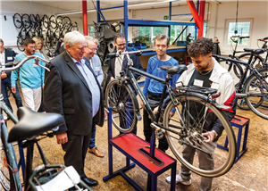 Minister Karl-Josef Laumann steht mit zwei Caritas-Vertretern in einer Fahrrad-Werkstatt des Schloss Bellinghoven und schaut Auszubildenden bei der Arbeit zu