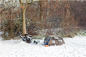 Der Schlafplatz eines Obdachlosen mit aufgebautem Zelt, Fahrrad und Einkaufkarre, der sich auf einer mit Schnee bedeckten Wiese vor einem Waldstück befindet