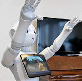 Der Roboter Pepper steht mit erhobenen Armen in einem Gemeinschaftsraum. Im Hintergrund ist ein Holzschrank mit Fernseher zu sehen.