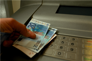 Eine Hand die einen 10- und zwei 20-Euroscheine zwischen den Finger hält und auf einem Geldautomaten aufliegt, von dem der untere Teil mit dem Tastenfeld zusehen ist