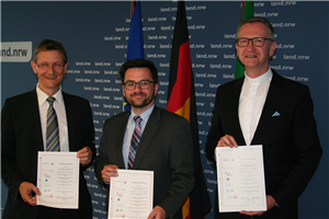 Dr. Frank Joh. Hensel, Thomas Kutschaty und Pfarrer Dr. Antonius Hamers stehen vor einer blauen Stellwand und halten drei Exemplare der Rahmenvereinbarung hoch.