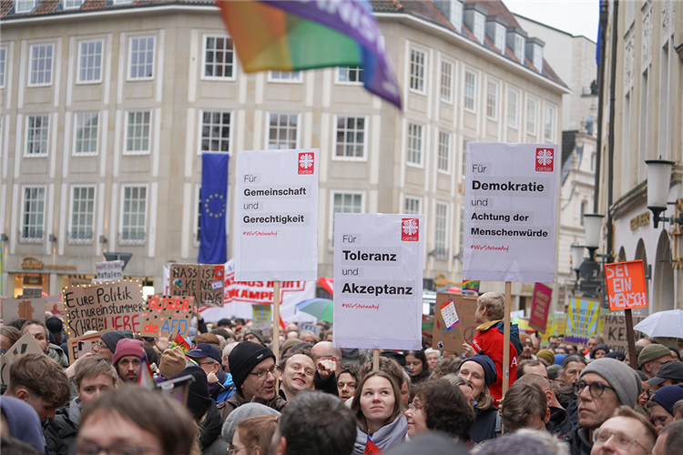 Eine Demonstration für Demokratie und Menschenwürde, die auf dem Prinzipalmarkt in Münster stattfindet. Die teilnehmenden Caritas-Mitarbeitenden halten drei Schilder hoch.