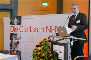 Diözesan-Caritasdirektor Burkard Schröders steht vor einem Pult mit einem Laptop, das neben einem Display der Caritas in NRW steht, und hält eine Rede