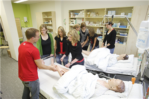 Sieben Pflegeschülerinnen und ein Pflegeschüler stehen an zwei Krankenbetten und führen Übungen an zwei Puppen durch