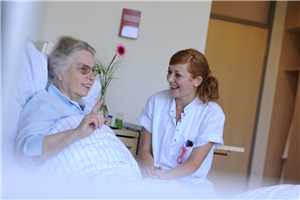 Eine Seniorin liegt in einem Krankenbett, eine Pflegerin sitzt daneben. Beide reden miteinander und lächeln.
