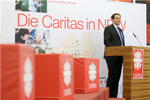 NRW-Staatssekretär Nathanael Liminski steht beim Ruhrgebietstag vor einem Rednerpult und hält sein Grußwort. Es sind außerdem ein Faltdisplay der Caritas in NRW und drei rote Caritas-Würfel zu sehen.