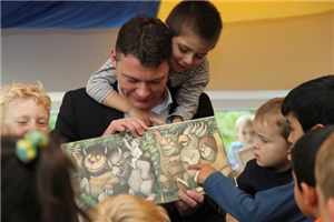 Dr. Jörg Geerlings (CDU) hält ein aufgeschlagenes Bilderbuch in der Hand und zeigt es einer Gruppe von Kindern. Einer der Jungen hängt ihm über den Schultern.