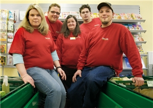 Ein Gruppenfoto von zwei Mitarbeiterinnen und drei Mitarbeitern von „Unser Laden – Lebensmittel & mehr“, die vor einem Zeitschriftenregal zwischen Warenkisten stehen