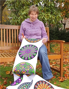 Petra Asch sitzt auf einer Holzbank und hält Buntstifte sowie ein langes Banner mit verschiedenen, bunten Mandalas in den Händen