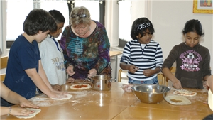 Eine Frau backt mit einer Gruppe Kinder gemeinsam Pizzen an einem großen Tisch