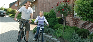 Ein Vater und seine Tochter sitzen auf ihren Fahrrädern mit einem Fuß auf dem Boden vor einem Wohnhaus