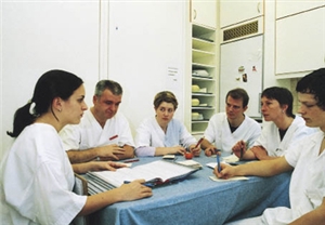Eine Besprechung mit sechs Pflegekräften, die dabei an einem Tisch in einem Personalraum sitzen.