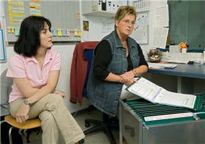 Zwei weibliche Pflegekräfte sitzen nebeneinander in einem Büro. Vor der rechten Pflegekraft steht ein Hängeregister mit einer daraufliegenden, aufgeklappten Akte.