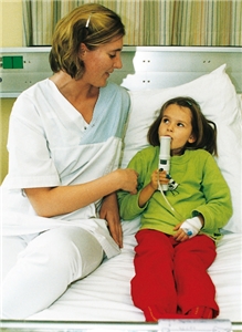 Eine Krankenschwester sitzt am Krankenbett eines Mädchens. Das Mädchen hält sich ein medizinisches Gerät an den Mund.