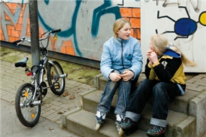 Zwei Mädchen reden, sitzend auf einer Treppen, miteinander. Im Hintergrund befindet sich eine mit Graffitis beschmierte Wand, links neben der Treppe steht ein Fahrrad an einer Laterne.