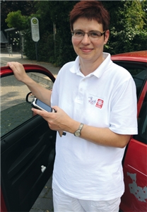 Christiane Gossmann steht ziwschen der geöffneten Tür ihres roten Pflegeautos und hat ein Handy in der Hand