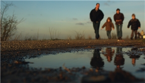 Eine Familie mit zwei Kindern beim Spaziergang über einen Wanderweg. Das Foto wurde vom Boden aus mit Fokus auf eine im Vordergrund liegende Regenpfütze aufgenommen.