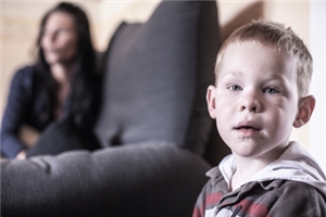 Ein Junge schaut an der rechten Bildseite in die Kamera, links daneben sitzt seine Mutter auf einem Sofa