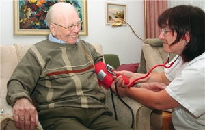 Eine Altenpflegerin misst bei einem, im Sessel sitzenden, Patienten den Blutdruck
