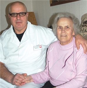 Schemsi Sadriu sitzt neben der Patientin Helga Ape, hält ihre Hand und legt seinen Arm über ihre Schulter