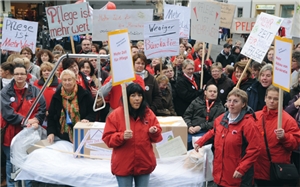Die Teilnehmer einer Pflegedemonstration stehen mit Bannern und Schildern auf einer Straße. Zwischen den Demonstraten steht ein Pflegebett mit zwei, mit Aufklebern versehenen, Kartons darauf.