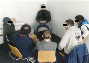 Ein Teilnehmer des Kurses sitzt in der Ecke eines Raumes. Fünf weitere Teilnehmer sitzen ihm viertelkreisförmig gegenüber bzw. umkreisen ihn.