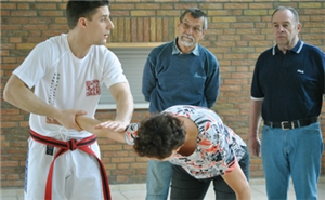 Ein Trainer demonstriert eine Selbstverteidigungstechnik an einer Senorin. Zwei weitere Senioren schauen dabei zu.