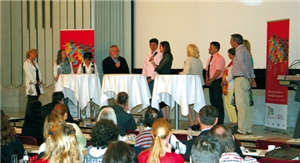 Im Hintergrund befindet sich eine Bühne mit drei Stehtischen und eine Personengruppe, zwei der Personen haben ein Mikrofon und reden miteinander. Im Vordergrund sitzt das Publikum der Veranstaltung.