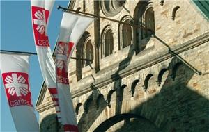 Der obere Teil der Frontseite eines Kirchengebäudes, aus dem drei Fahnenmasten mit Caritas-Fahnen hervorragen