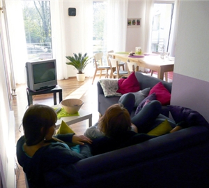 Zwei Frauen liegen zusammen auf einem Sofa. Der Blick der Kamera geht in den gesamten Wohnraum, in dem links ein Fernseher und rechts ein Esstisch stehen.