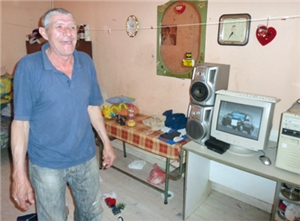 Ein Mann namens „Ein-Zahn“ steht lächelnd in seinem Wohnraum, in dem rechts ein Schreibtisch mit Computer und ein Abstelltisch zu sehen sind