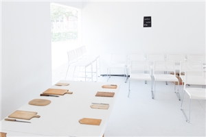 Installation: Ein komplett in weiß gehaltener Raum mit einem großen Fenster oben links. In dem Raum stehen viele weiße Stühle und Tische auf denen Holzbrettchen mit je einem Messer bereit liegen