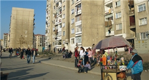 Eine Straße im Stadteil Stolipinovo (Plovdiv) auf der eine große Menschenmenge unterwegs ist. Im Hintergrund befinden sich alte Plattenbauten, vorne rechts steht ein Händler mit einem Verkaufswagen.