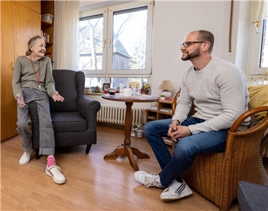 Eine ältere Bewohnerin des Wilhelm-Knappmann-Hauses sitzt auf der Armlehne eines Sessels in ihrem Zimmer; ihr gegenüber eine Caritas-Mitarbeiter.