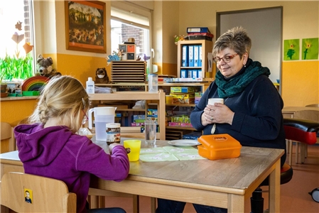 Eine Erzieherin sitzt mit einem Kind an einem Tisch im Kindergarten beim Frühstück.