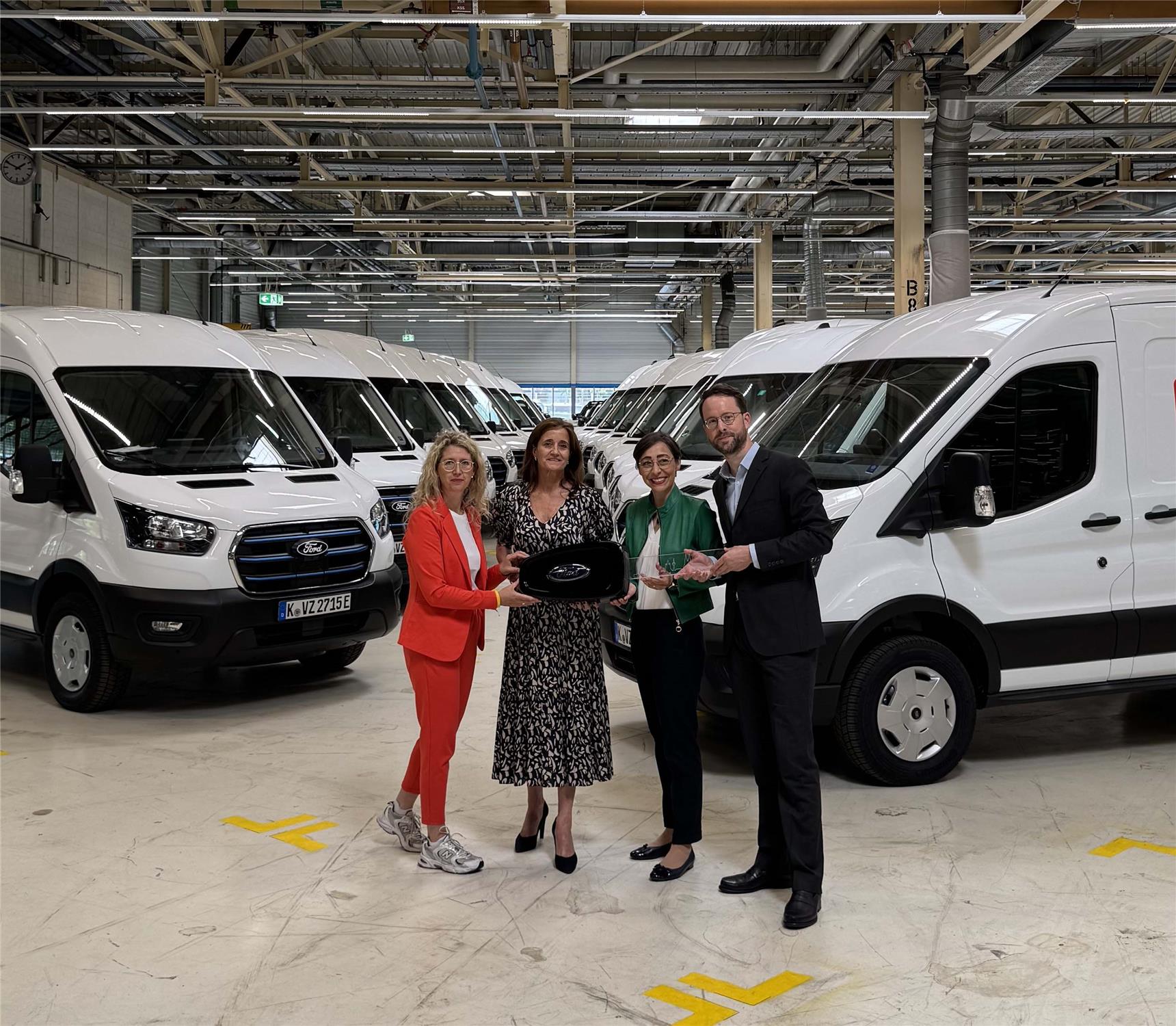 In einer Werkshalle parken in zwei Reihen zahlreiche weiße Ford-Transportfahrzeuge. Davor steht eine Gruppe mit Vertreterinnen und Vertretern von Ford und der Caritas anlässlich der offiziellen Fahrzeugübergabe.