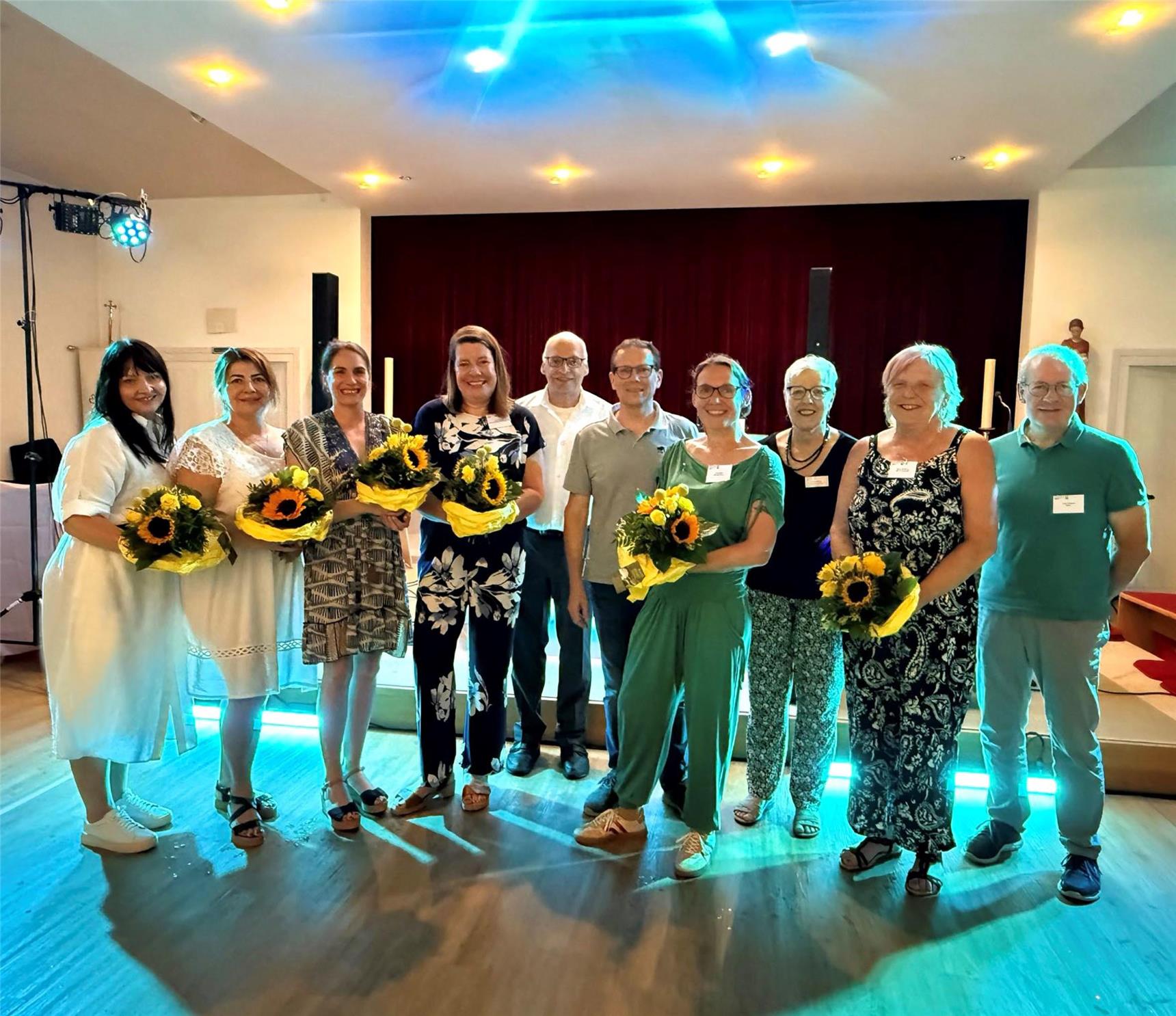Gruppenfoto, das zehn sommerlich-festlich gekleideten Menschen in einem Saal zeigt. Darunter sechs Frauen, die Blumensträuße mit Sonnenblumen in den Händen halten.