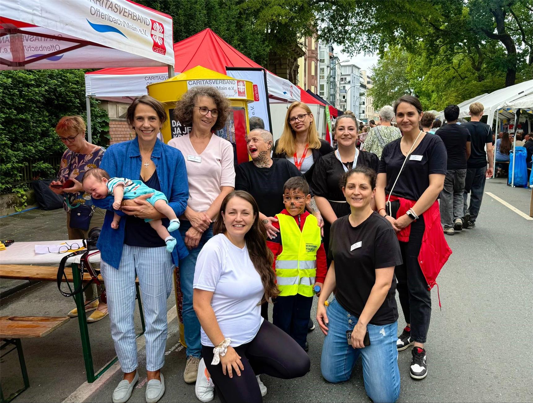 Gruppenbild von 8 Caritas-Mitarbeitenden und zwei Kindern. Sie stehen vor dem Caritas-Infostand auf der Festmeile an der Offenbacher Mainstraße.