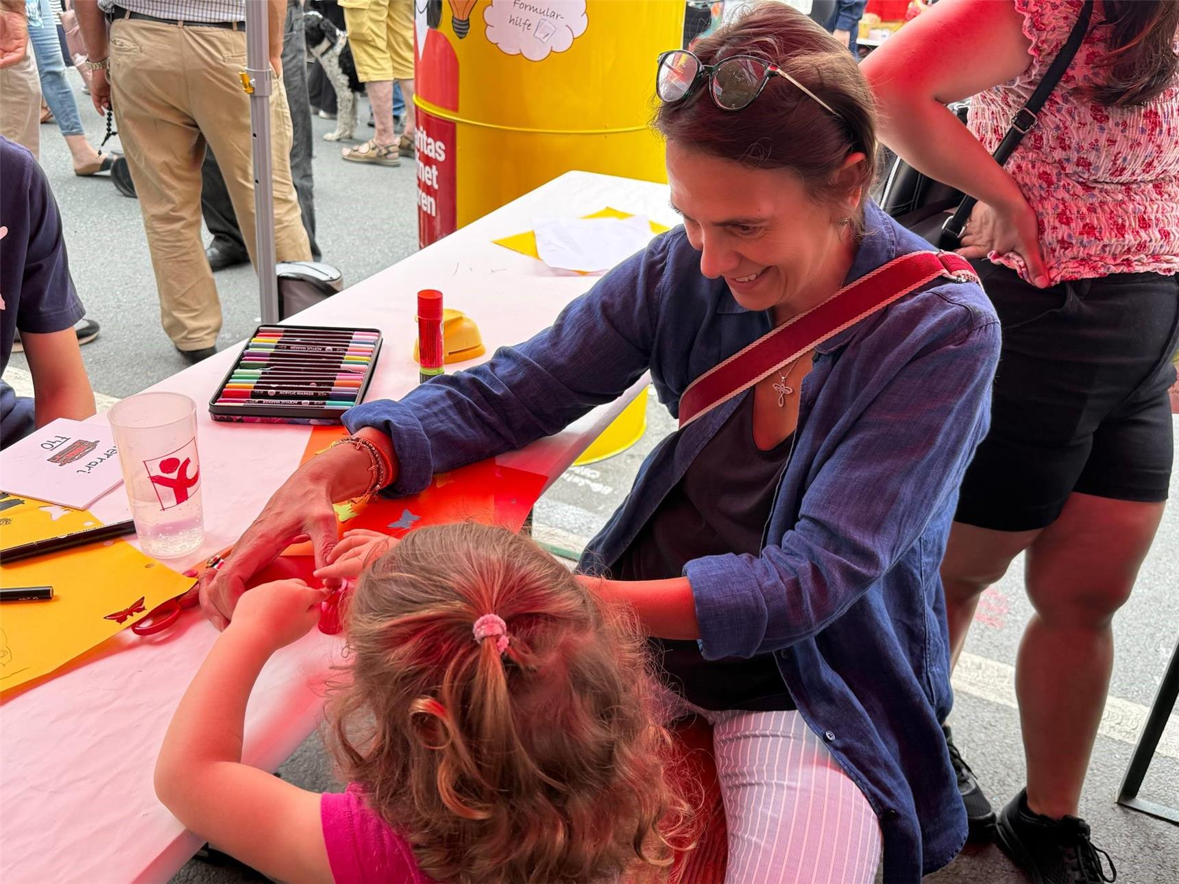 Ein kleines Mädchen sitzt am Tisch des Infostandes und bastelt. Caritas-Bereichsleiterin Anette Bacher hilft ihr dabei.