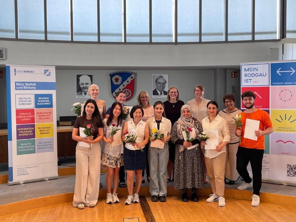 Gruppenfoto von acht Integrationslotsinnen und einem Integrationslotsen im Rathaus Jügesheim in Rodgau. Alle halten Blumen und eine Urkunde in den Händen. Ebenfalls auf dem Gruppenfoto in der hinteren Reihe zu sehen: Vertreterinnen der Stadt Rodgau und der Caritas.