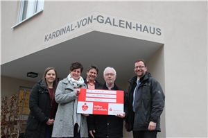 Fünf Personen mit einem überdimensionierten Scheck vor dem Eingang des Kardinal-von-Galen-Hauses