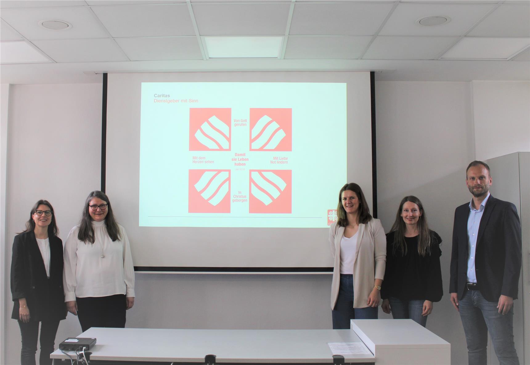 Fünf Personen stehen nebeneinander vor einer Leinwand in einem Konferenzraum und lächeln in die Kamera. Auf der Leinwand ist eine Präsentationsfolie der Caritas mit dem Titel „Dienstgeber mit Sinn“ zu sehen, die vier rote Felder mit weißen Symbolen und Texten enthält, darunter „Von Gott gerufen“, „Mit dem Herzen sehen“, „Mit Liebe Not lindern“, „In Christus geborgen“ sowie der zentrale Satz: „Damit sie Leben haben“ (Joh 10,10).