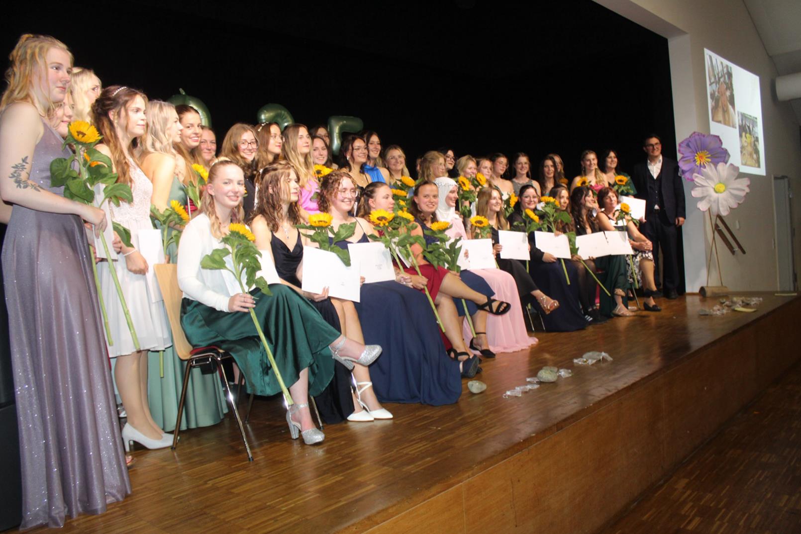 Viele Personen stehen auf einer Bühne. Diese ist dekoriert mit großen Blumen. Alle Absolvent:innen haben eine Sonnenblume in der Hand.