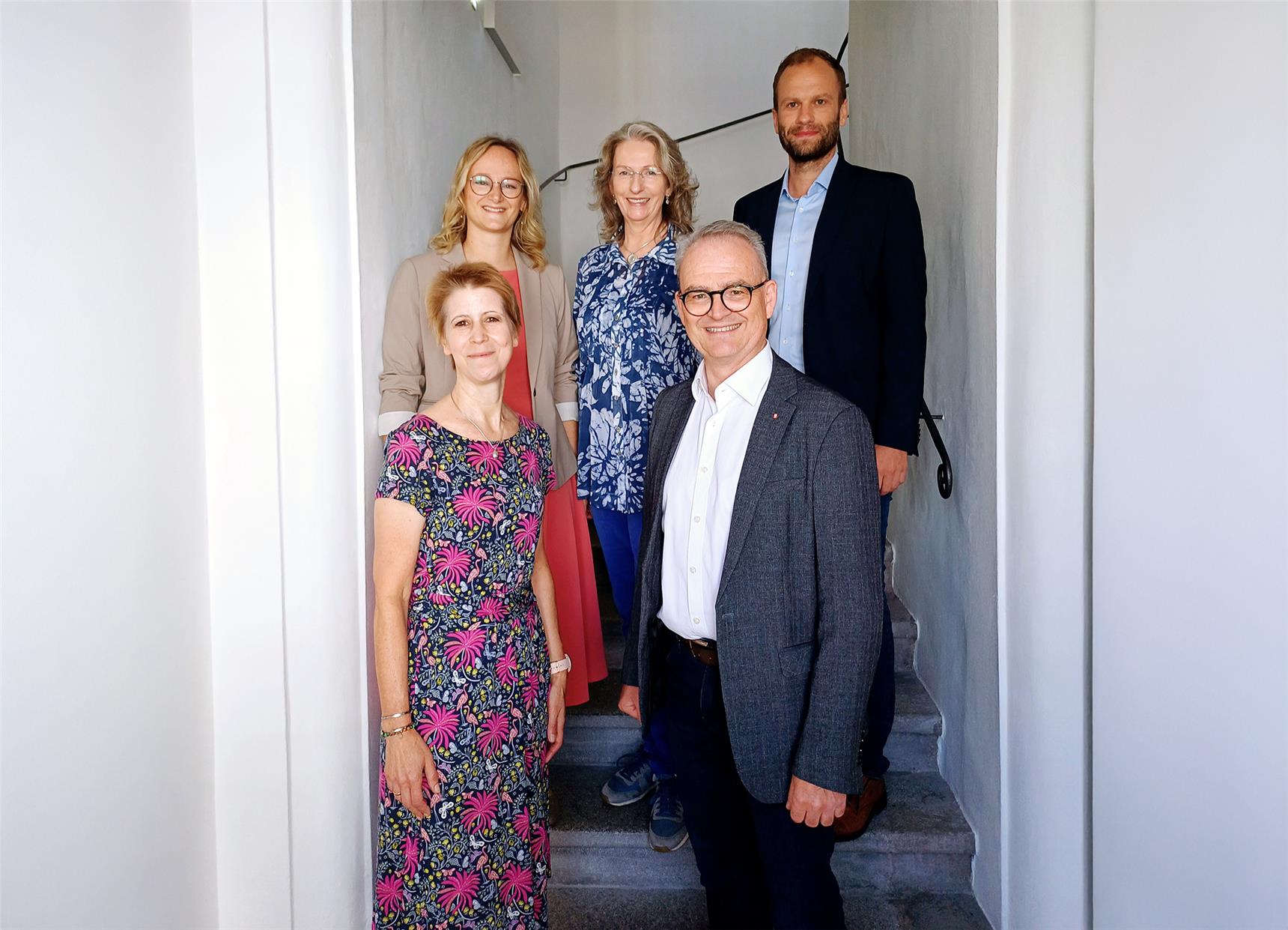 Fünf Personen stehen auf einer Treppe in einem hellen Gebäude und blicken freundlich in die Kamera. Zwei Männer im Anzug und drei Frauen in gemusterten Kleidern oder Blusen vermitteln einen professionellen, aber einladenden Eindruck – möglicherweise ein Team aus dem Gesundheits- oder Bildungsbereich.