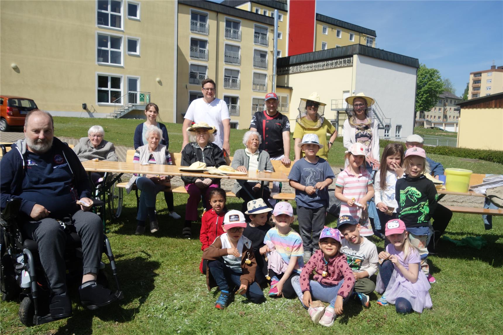 Mehrere Personen sitzen auf einer Bank und weitere stehen hinter der Bank. Kinder stehen und sitzen davor im Gras. Alle lachen in die Kamera.
