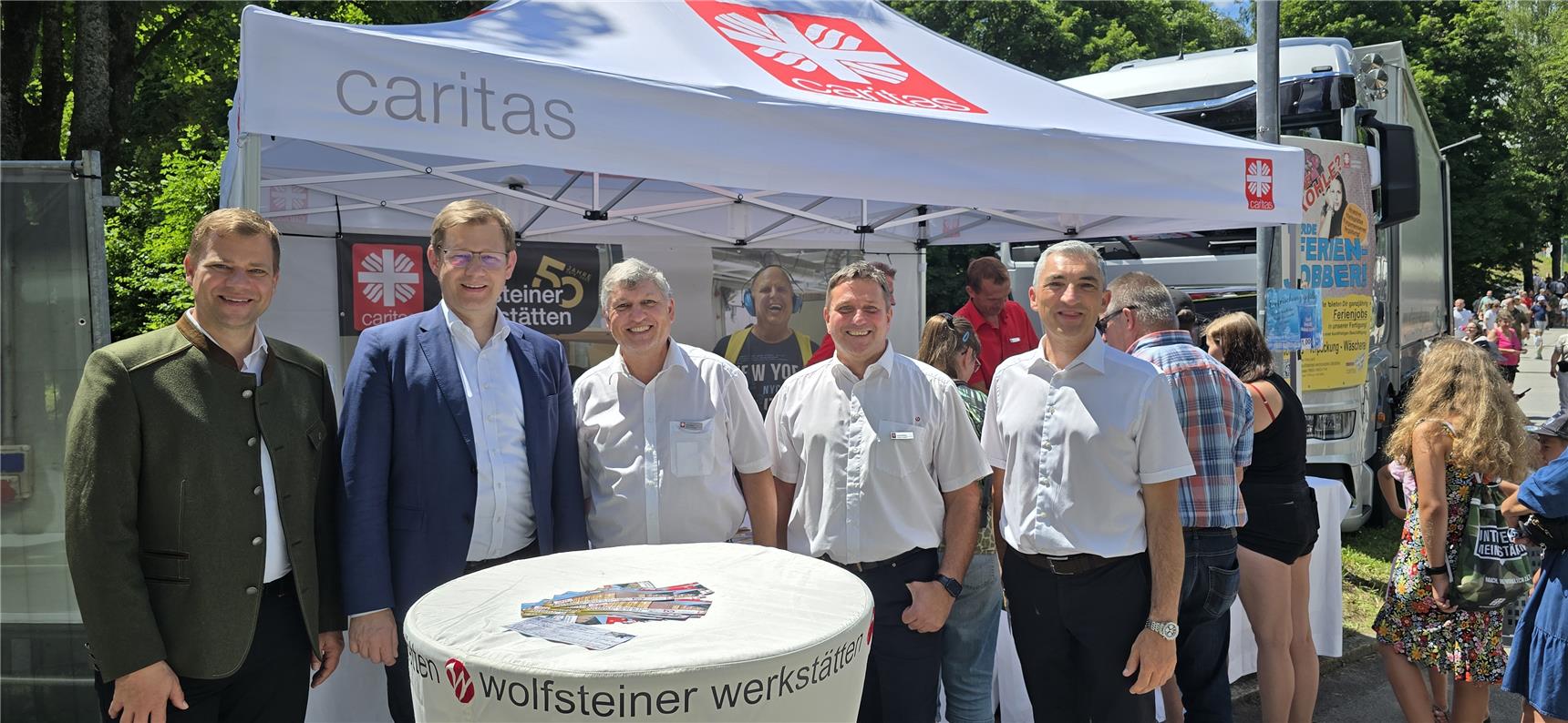 5 Personen stehen vor einem Caritas-Zelt und lachen in die Kamera. Im Zelt: Banner und Flyer. 