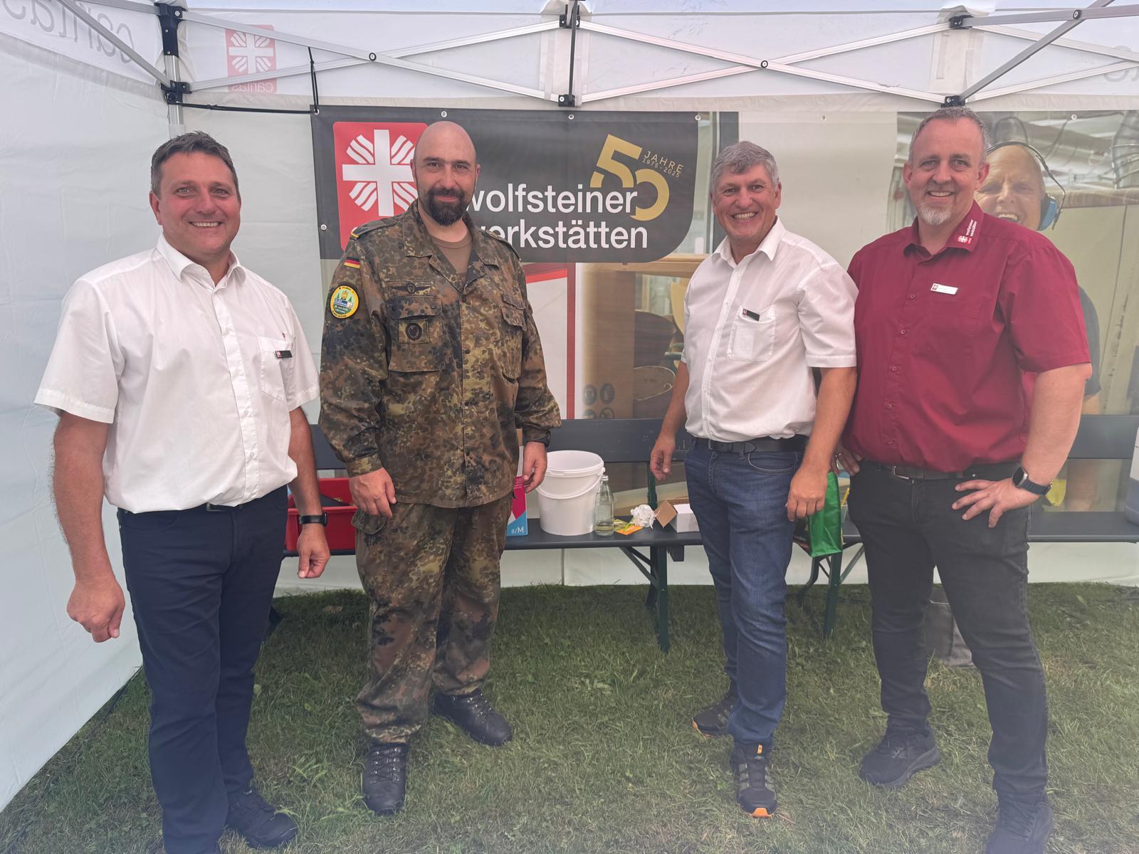 4 Personen stehen in einem Caritas-Zelt und lachen in die Kamera. Eine Person davon trägt eine Uniform. Im Hintergrund ist ein Banner der Caritas mit 