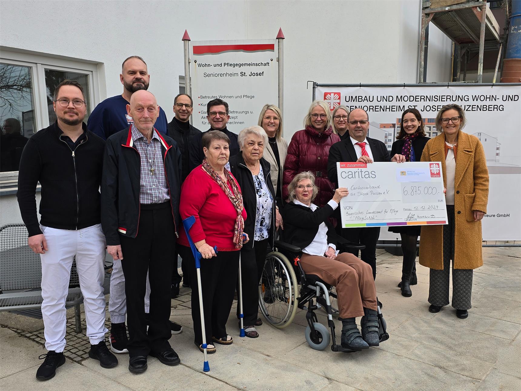 Das Bild zeigt eine Gruppe von Menschen vor einem Gebäude mit einem Schild der "Wohn- und Pflegegemeinschaft Seniorenheim St. Josef." Die Gruppe besteht aus Senioren, Pflegekräften und Vertretern, die