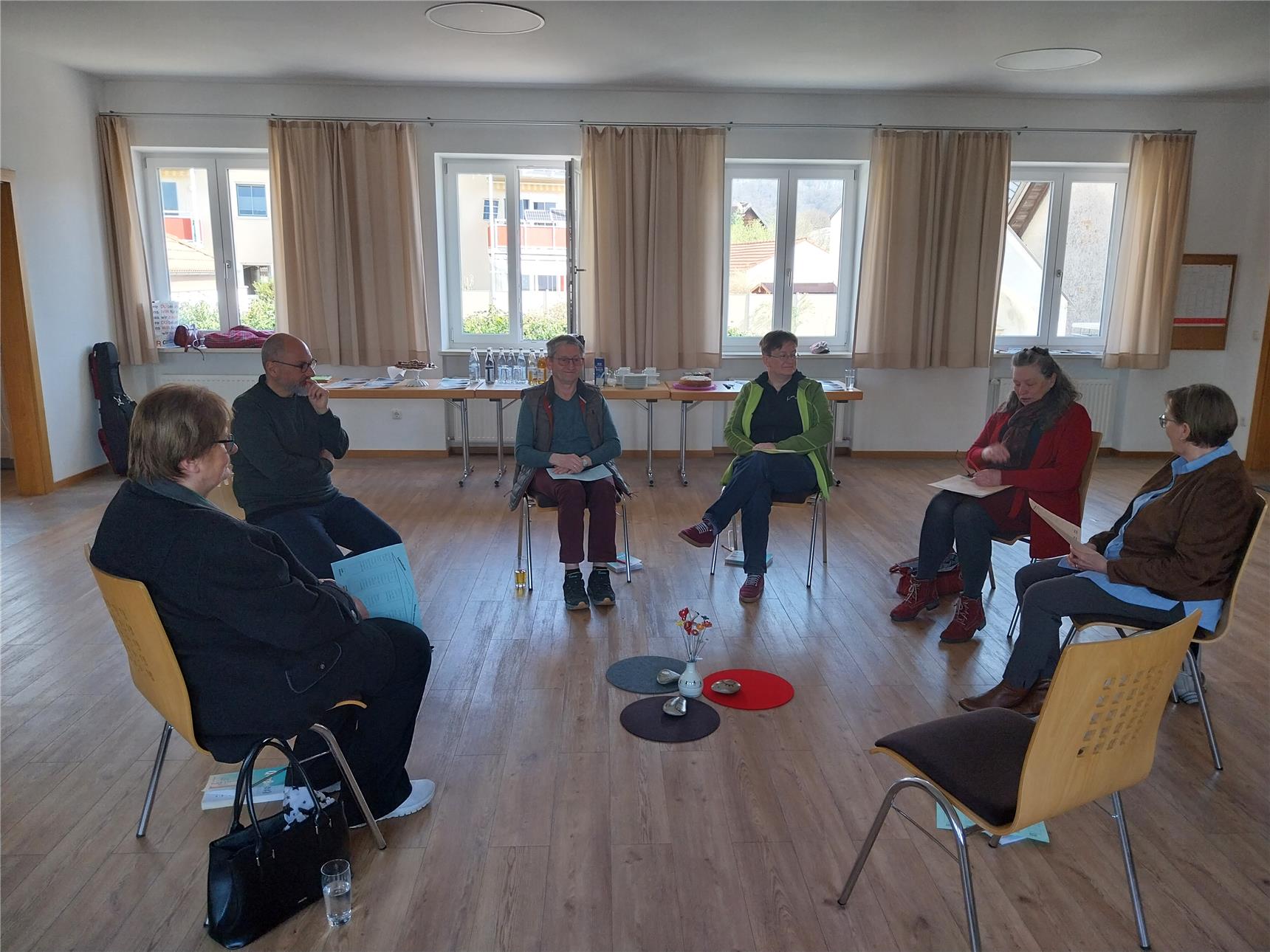 Sechs Personen sitzen im Kreis in einem hellen Raum und führen ein Gespräch oder eine Gruppensitzung. In der Mitte stehen kleine Deko-Objekte auf runden Filzunterlagen.
